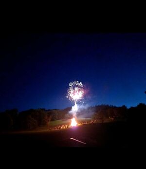 Feuerwerk erhellt den Nachthimmel über einer feiernden Menschenmenge neben einem großen Feuer.